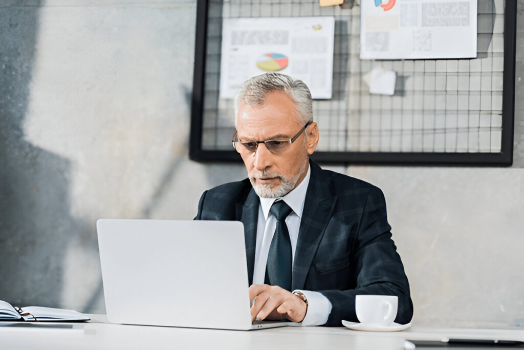 Elderly businessman in a suit working on a laptop in office.