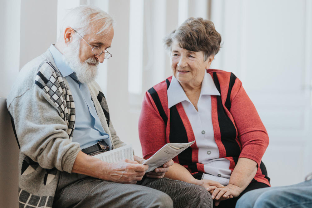 Elderly couple sitting together, reading newspaper and smiling warmly.