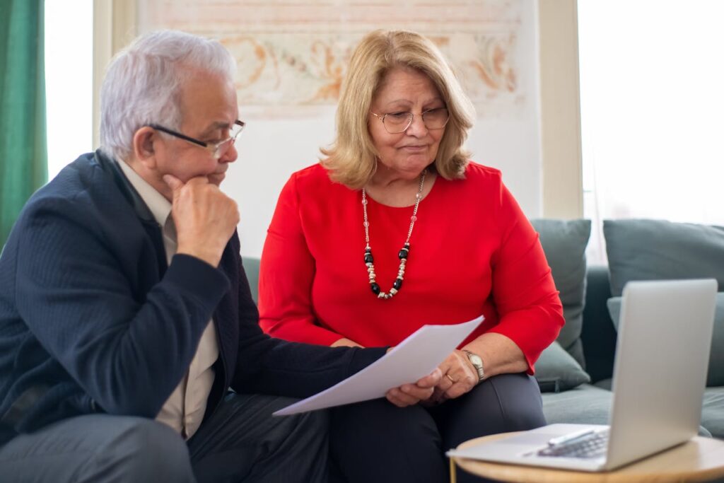 Elderly couple sitting on sofa, reviewing documents together on laptop.