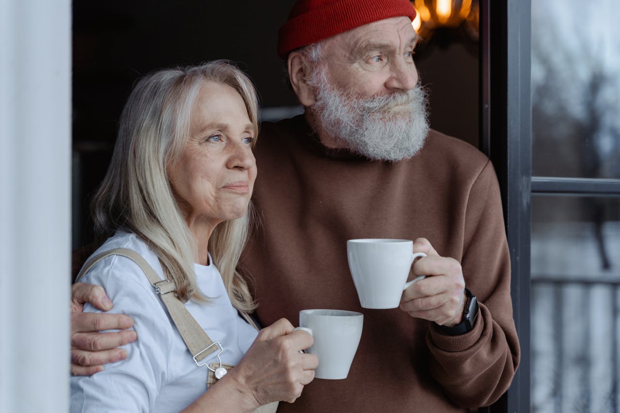 Elderly couple standing together with coffee cups, looking outside.