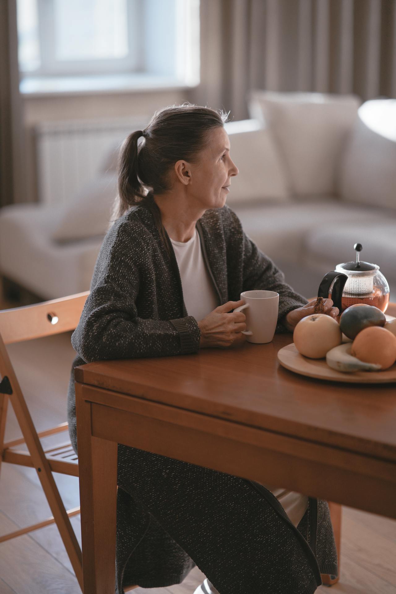 Older woman sitting at wooden table, holding cup, looking thoughtful.